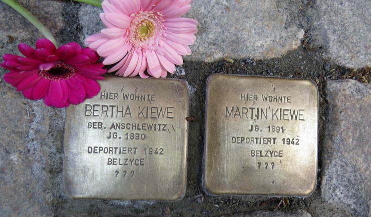 zwei Metallplatten mit Inschrift im Boden inmitten von Pflastersteinen eingelassen, daneben zwei rote Blumen