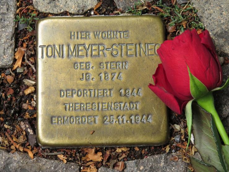 Metallplatte mit Inschrift im Boden inmitten von Pflastersteinen eingelassen, daneben eine rote Rose