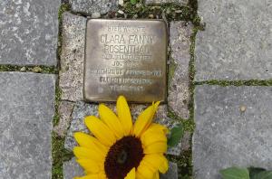 Metallplatte mit Inschrift im Boden inmitten von Pflastersteinen eingelassen, daneben eine Sonnenblume