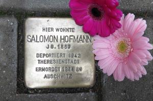 Metallplatte mit Inschrift im Boden inmitten von Pflastersteinen eingelassen, daneben zwei rote Gerbera