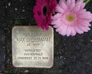 Metallplatte mit Inschrift im Boden inmitten von Pflastersteinen eingelassen, daneben zwei rote Gerbera