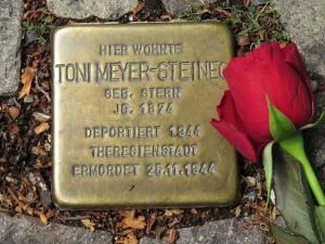 Metallplatte mit Inschrift im Boden inmitten von Pflastersteinen eingelassen, daneben eine rote Rose